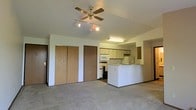 This single-floor space showcases an open-concept layout that seamlessly connects the living area to the kitchen, creating a functional and cohesive environment. The living area is finished with wall-to-wall carpeting that lends a comfortable, understated feel, while a ceiling fan with an integrated light fixture and an accompanying track light provide both ventilation and overhead illumination. Along one wall, two wood-paneled sliding closet doors offer generous built-in storage, accompanied by an additional solid-panel door providing access to adjoining areas. The kitchen is thoughtfully designed with light-toned cabinetry spanning the upper and lower sections, offering ample storage space. A white range, microwave, and sink are neatly arranged along the kitchen wall, while a white peninsula counter extends outward, creating a natural divide between the kitchen and living areas and providing additional workspace or casual seating potential. Recessed under-cabinet fluorescent lighting brightens the kitchen workspace. The overall color palette — soft neutrals throughout the walls, cabinetry, and flooring — contributes to a clean, bright, and practical living environment well-suited for everyday use.