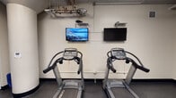 Located at 2112 S Michigan Ave in Chicago's South Loop neighborhood, this fitness center is a single-floor facility designed to provide a functional and well-equipped workout environment. The space features neutral-toned walls, dark rubberized flooring, and exposed ceiling infrastructure, giving it a utilitarian yet organized aesthetic. Structural columns are integrated throughout, adding an architectural element to the interior layout.

The cardio area is anchored by two Cybex treadmills positioned side by side, each paired with wall-mounted flat-screen televisions overhead for entertainment during workouts. A dedicated free weights zone includes a full dumbbell rack with a range of weights, exercise mats, and medicine balls, complemented by a large wall mirror that enhances the sense of space and aids in form checking. Strength training equipment, including a cable machine, is also available within the facility.

An Aquafina beverage vending machine provides convenient hydration access, while a snack and supply display cabinet near the entrance offers additional refreshments and essentials. A hand sanitizer station and waste bin are positioned near the entry door, promoting cleanliness. The overall layout is efficiently organized, making the most of the available space for a variety of workout routines.