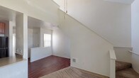 Located at 420 Activity in Oceanside, this two-story unit offers a well-considered layout with distinct living zones spread across both levels. The main living area immediately draws the eye upward with dramatically vaulted ceilings that lend an open, airy quality to the space. A ceiling fan is positioned overhead, and the room transitions between plush carpeting and rich, dark hardwood-style flooring, creating visual contrast throughout.

The kitchen is accessible via a pass-through window opening from the living area, providing a connected yet defined space. Inside, the kitchen is equipped with dark-toned cabinetry, black appliances including a range, microwave, and refrigerator, and countertops with a sink along the perimeter. The dark flooring continues here, complementing the warm wood tones of the cabinetry.

A staircase leads to the upper level, where a loft-style space features sloped ceilings that follow the roofline, adding architectural character. Carpeting covers this level, and an open railing overlooks the lower floor. Additional rooms include a carpeted bedroom with clean white walls and a full bathroom featuring a tub-shower combination, a single vanity with granite-style countertop, and a large mirror. Natural light filters in through windows with vertical blinds throughout the unit.
