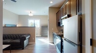 Located at 2675 Sycamore Rd in DeKalb, Illinois, this single-floor unit offers a well-designed, open-concept layout that seamlessly connects the kitchen, living, and sleeping areas. The kitchen is appointed with rich, dark-stained cabinetry, granite countertops featuring a bold veining pattern, a stone-accented backsplash, and stainless steel appliances including a refrigerator, dishwasher, and microwave. Light wood-tone flooring runs throughout, enhancing the bright and airy feel of the space, which benefits from natural light streaming through well-placed windows. The living area provides a relaxed setting with warm neutral-toned walls and comfortable seating, including a tufted leather sofa and an accent chair arranged around a coffee table. A low decorative ledge with subtle lighting adds architectural interest to the living space. The bedroom is a calm retreat, featuring coordinated dark-finish furniture and elegant wall-mounted sconces flanking the headboard, creating a cozy and restful ambiance. The bathroom is finished with large-format marble-look tile on both the walls and shower surround, a glass-enclosed walk-in shower with oil-rubbed bronze fixtures, and granite countertops, delivering a clean and refined aesthetic throughout.