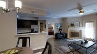 This thoughtfully designed single-floor residence showcases an open-concept floor plan that seamlessly connects the main living areas. The kitchen is outfitted with dark espresso cabinetry, stainless steel appliances including a French door refrigerator, and a white breakfast bar with seating — ideal for casual dining or entertaining. Rich, dark hardwood-style flooring flows throughout, anchoring the space with warmth and contrast against the soft neutral wall tones and crisp white crown molding that accents the ceilings.

The living area is centered around a classic white fireplace with an ornate decorative screen, adding a traditional focal point to the room. A media console and area rug define the lounge space, while the open sightlines contribute to an airy, spacious feel. Natural light filters through the windows, enhancing the bright and welcoming ambiance throughout.

Through a wide doorway, the bedroom offers a cozy retreat with warm lighting, a built-in shelving unit, and ample room for furnishings. The cohesive design language — blending contemporary finishes with classic architectural details — carries through every area of the home, creating a polished and comfortable living environment.