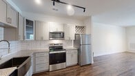 This well-appointed one-bedroom, one-bathroom unit in Sneads Ferry, NC offers a thoughtfully designed single-floor layout with modern finishes throughout. The open-concept living, dining, and kitchen area serves as the heart of the home, featuring rich hardwood-style flooring, track lighting, and large windows that invite abundant natural light and outdoor views. The kitchen is equipped with stainless steel appliances — including a range, over-the-range microwave, refrigerator, and dishwasher with a stainless farmhouse-style sink — complemented by white shaker cabinetry, granite countertops, a subway tile backsplash, and a built-in wine rack. The bedroom is finished with plush carpeting, a ceiling fan, and dual windows overlooking a pool area. The bathroom features hardwood-style flooring, a full tub/shower combination, granite vanity, and grey cabinetry. A dedicated utility room houses a full-size washer and dryer with overhead wire shelving for added storage. A walk-in closet with open wood shelving provides generous organization space, rounding out this clean, contemporary unit.