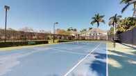 This outdoor recreational facility centers around a full-sized, hard-surface tennis court finished in a classic blue and green color scheme. The court is equipped with a regulation net and is enclosed by a perimeter chain-link fence, ensuring a safe and contained playing environment. Overhead court lighting is provided by a tall pole-mounted fixture, allowing for extended play beyond daylight hours. A green bench is positioned along the courtside, offering a convenient resting area for players and spectators. The surrounding landscape features mature palm trees, deciduous trees, and well-kept greenery that frame the court with a tropical, resort-like atmosphere. The facility benefits from abundant natural sunlight under an open, clear blue sky, reflecting a warm and inviting climate. In the background, well-maintained residential buildings with light-colored facades are visible beyond the fence line, suggesting the court is part of a broader residential community or complex. The overall setting is clean, well-maintained, and designed to support an active outdoor lifestyle.