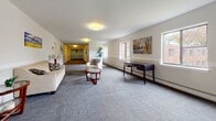 Located at 905 Burnside Ave in East Hartford, Connecticut, this single-floor communal lounge space offers a well-appointed gathering area designed for comfort and functionality. The open layout is finished with patterned, tile-cut carpeting in neutral grey tones that extends throughout, providing a clean and cohesive aesthetic underfoot.

The space is bathed in natural light through multiple large windows that line the walls, offering views of the surrounding landscaped exterior. Baseboard heating runs along the perimeter, ensuring year-round comfort. The neutral-toned walls are accented with a wainscoting-style horizontal rail detail, lending a touch of classic architectural character to the interior.

Seating is thoughtfully arranged across distinct zones: a casual lounge area features a cream-colored sofa and matching armchair centered around a glass-top coffee table, while a secondary seating nook showcases dark leather club chairs paired with a wooden side table. Decorative indoor plants, framed landscape artwork, and fresh floral accents contribute warmth and visual interest throughout.

A writing desk along one wall provides a functional workspace, and a well-lit corridor leads to additional areas of the building. The overall ambiance is calm, clean, and welcoming — well-suited for a residential common area or small community gathering space.