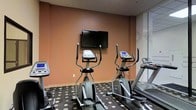 This fitness center is a dedicated, single-floor exercise space designed to support a well-rounded cardio workout routine. The room is outfitted with multiple stationary bikes, elliptical trainers, and treadmills — including Matrix-brand equipment — providing users with a solid range of aerobic exercise options. A flat-screen television is wall-mounted at a comfortable viewing height, allowing guests to stay entertained during their workouts. The warm, earthy tones of the walls create an energizing yet approachable atmosphere, complemented by the distinctive patterned carpet featuring a polka-dot design that adds a touch of character to the space. The layout is thoughtfully arranged to allow comfortable movement between machines without feeling cramped. Large mirrored panels along one wall enhance the sense of openness and allow users to monitor their form during exercise. The overall ambiance is clean, functional, and well-maintained, reflecting a facility intended to serve guests seeking a convenient and reliable workout experience. The space is straightforward in its purpose, prioritizing practical fitness amenities in a comfortable and accessible environment.