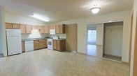 This single-floor residence offers a practical and well-organized layout designed for comfortable everyday living. The kitchen serves as a central feature of the space, outfitted with light wood cabinetry that provides generous storage both above and below the countertops. Dark laminate counters contrast neatly with white appliances, including a freestanding range with oven, a built-in microwave, and a dishwasher. A double-basin sink sits beneath a fluorescent light fixture, ensuring a well-lit workspace. The kitchen floor is finished in neutral-toned tile, lending a clean and cohesive look to the area. A full-height pantry or utility door adds further storage convenience adjacent to the kitchen. The space opens seamlessly through a sliding glass door into a carpeted room beyond, which benefits from a bright window that draws in natural light, creating an airy and open atmosphere. The overall palette of soft creams and warm wood tones gives the space a welcoming, understated character. The single-floor layout ensures easy accessibility and a straightforward flow between areas, making it a functional and comfortable living environment.