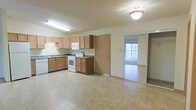 This single-floor residence offers a practical and well-organized layout designed for comfortable everyday living. The kitchen serves as a central feature of the space, outfitted with light wood cabinetry that provides generous storage both above and below the countertops. Dark laminate counters contrast neatly with white appliances, including a freestanding range with oven, a built-in microwave, and a dishwasher. A double-basin sink sits beneath a fluorescent light fixture, ensuring a well-lit workspace. The kitchen floor is finished in neutral-toned tile, lending a clean and cohesive look to the area. A full-height pantry or utility door adds further storage convenience adjacent to the kitchen. The space opens seamlessly through a sliding glass door into a carpeted room beyond, which benefits from a bright window that draws in natural light, creating an airy and open atmosphere. The overall palette of soft creams and warm wood tones gives the space a welcoming, understated character. The single-floor layout ensures easy accessibility and a straightforward flow between areas, making it a functional and comfortable living environment.