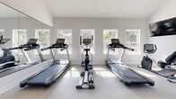 Located at 11909 Apache Ave in Savannah, Georgia, this single-floor fitness center is designed to provide a comprehensive and comfortable workout environment. The space is defined by its soaring vaulted ceilings, which lend an airy, open feel throughout. Crisp white walls and neutral carpeting create a clean, bright aesthetic that enhances the sense of spaciousness.

The facility is divided into two primary workout zones. The cardio area features multiple treadmills and an elliptical machine positioned in front of a generous row of windows that frame views of the surrounding outdoor pool area and lush Georgia landscape, allowing natural light to flood the space. A large floor-to-ceiling mirror wall runs along one side, visually expanding the room and providing functional visibility during workouts.

The strength training zone is equally well-appointed, housing a multi-function cable and weight machine, a workout bench, and a tiered dumbbell rack stocked with a full range of free weights. Additional windows along this section of the facility overlook neighboring grounds, maintaining the bright and open atmosphere throughout.

A wall-mounted flat-screen television and ceiling fan add convenience and comfort. The overall design balances functionality with a clean, modern aesthetic, making this fitness center a well-rounded amenity for residents of the community.