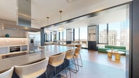 Located at 10-14 LeCount Place in New Rochelle, NY, this sophisticated amenity lounge offers a thoughtfully designed, single-floor layout that seamlessly blends a fully equipped kitchen with open dining and social gathering spaces. The kitchen is a standout feature, appointed with stainless steel appliances including wall ovens and a dishwasher, sleek white cabinetry, stone-tiled backsplash, and a generously sized island clad in blue mosaic tile topped with a quartz counter. Globe pendant lights with brass accents illuminate the space with a warm, contemporary glow. The open dining and lounge area transitions effortlessly from the kitchen, showcasing a stunning herringbone hardwood floor and multiple seating arrangements with modern chairs and tables suited for both casual dining and collaborative gathering. Floor-to-ceiling windows line the perimeter, flooding the space with abundant natural light and offering expansive panoramic views of the New Rochelle skyline and beyond. A cozy lounge nook features a bold emerald green sectional sofa set against decorative botanical wallpaper, providing an inviting retreat. The overall ambiance is modern and upscale, with a refined palette of dark steel framing, natural wood tones, and contemporary furnishings that create an energetic yet comfortable communal environment.