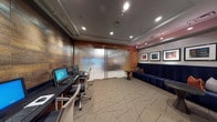 This single-floor business center is thoughtfully divided into two functional zones, each with its own distinct character while sharing a cohesive design language. The workstation area lines one wall with a sleek dark desk surface accommodating multiple desktop computers and ergonomic task chairs. A striking textured wall panel in warm, earthy tones with varied brown hues serves as a dramatic backdrop, illuminated by directional track lighting that highlights its rich surface detail. Frosted sliding glass panels with a rippled texture separate this zone, providing privacy while maintaining an open, modern aesthetic.

The adjacent lounge area features a substantial navy blue upholstered bench sofa accented with rust-orange throw pillows, offering a comfortable waiting or collaboration space. A series of four framed artworks with vibrant, colorful compositions are displayed above the sofa, spotlit by track lighting mounted on the ceiling. Small round side tables in dark wood flank the seating. The space is anchored throughout by geometric-patterned carpeting in neutral tones. A secondary alcove contains built-in warm wood cabinetry, additional office equipment, and further framed artwork, completing this well-appointed and professional environment.