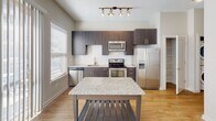 Located at 616 Oberlin Rd in Raleigh, NC, this single-floor unit offers a clean, contemporary design with a cohesive neutral palette throughout. The kitchen serves as a standout feature, outfitted with dark espresso cabinetry, a white subway tile backsplash, granite countertops, and a full suite of stainless steel appliances including a dishwasher, range, microwave, and refrigerator with ice maker. A granite-topped island with a slatted base provides additional prep space and anchors the open connection to the living area, which features hardwood-style flooring, a ceiling fan, and a recessed upper section that adds architectural interest to the ceiling line. A hallway with louvered closet doors and overhead ventilation leads to the private areas of the unit, including a carpeted bedroom with a unique clerestory window detail that draws in natural light from an elevated angle. The bathroom is well-appointed with a dark wood vanity, granite countertop, a large mirror, and a full tub-and-shower combination, all set on warm hardwood-style flooring. Throughout the unit, white trim, soft neutral walls, and thoughtful lighting create a bright and functional living environment.