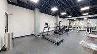 Located at 1000 Washington Ave in St. Louis, Missouri, this fitness center offers a well-equipped, single-floor layout designed to support a comprehensive workout experience. The main exercise area is generously sized, featuring durable rubber flooring throughout that provides both safety and comfort underfoot. The space is outfitted with a robust selection of True-brand cardio machines, including treadmills and recumbent bikes with interactive digital displays, alongside a cable machine and a variety of strength-training stations with adjustable benches. A large wall-mounted mirror along one side of the main floor visually expands the space and allows users to monitor their form during workouts. Structural columns are integrated tastefully into the layout, lending an open, loft-like character to the interior. The black drop-panel ceiling with recessed panel lighting creates a sleek, modern aesthetic while providing even, bright illumination throughout. A stainless steel dual drinking fountain is conveniently positioned near the bathroom entrance. The bathroom provides essential facilities to complete the experience. The overall ambiance is clean, contemporary, and functional, making this fitness center a practical and well-appointed amenity.