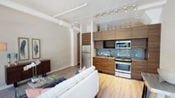 This single-floor loft-style space showcases a thoughtfully designed open-concept layout that seamlessly connects the living and kitchen areas. The design draws on industrial influences, most notably through the exposed white ductwork and structural columns that run along the ceiling, adding character and an urban edge to the interior.

The kitchen is a standout feature, outfitted with rich wood-grain cabinetry spanning floor to ceiling and complemented by a striking teal and blue mosaic tile backsplash. Stainless steel appliances — including a refrigerator, range, and built-in microwave — lend a clean, modern aesthetic. Track lighting mounted along the ceiling provides targeted illumination throughout the kitchen and living areas.

The living space flows naturally from the kitchen, anchored by light-toned hardwood flooring that runs throughout, bringing warmth and continuity to the interior. A white upholstered sofa and glass coffee table furnish the lounge area, creating an inviting and contemporary atmosphere.

Generous ceiling height enhances the sense of openness, while the overall design balances industrial rawness with modern refinement. The cohesive use of warm wood tones, metallic accents, and bold tile work gives this space a distinctive and well-curated character.