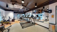 Located at 8 Warner St in Savannah, GA, this fitness facility is thoughtfully designed to accommodate a full range of workout disciplines within a single-floor layout. The space features a dark, industrial-style ceiling with exposed mechanical systems and recessed track lighting, creating a modern, energetic atmosphere. White walls contrast sharply with the dark ceiling, while a combination of gray and orange rubber flooring defines distinct workout zones throughout the facility.

The cardio area is anchored by a row of TRUE-brand treadmills equipped with individual digital screens, complemented by rowing machines, a stair climber, and additional aerobic equipment. Full-length mirrors along one wall visually expand the space and allow users to monitor their form. Large windows along the perimeter allow natural light to filter in while maintaining privacy.

The strength training zone includes a multi-functional cable machine, a rack of kettlebells, dumbbells in varying weights, battle ropes, a power rack, and an adjustable bench—offering comprehensive free-weight and resistance training options. A dedicated functional training area features a highlighted orange rubber floor section ideal for dynamic movement exercises.

A frosted glass partition wall with a glass door separates zones within the facility, adding a polished architectural detail. Ceiling-mounted HVAC units ensure climate control throughout. The overall design balances industrial aesthetics with practical, user-focused functionality.