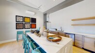 This single-floor space in Modesto, California showcases a thoughtfully designed, modern kitchen with an open and airy layout. The centerpiece is a generously sized island topped with sleek quartz countertops, lined with vibrant teal bar stools that add a bold pop of color against the neutral palette of light gray walls and pale hardwood flooring.

The kitchen is equipped with stainless steel appliances, including a dishwasher and undercounter refrigerator units, complemented by warm wood-toned cabinetry along the perimeter wall. Floating natural wood shelves provide open storage above the counters, contributing to the clean, minimalist aesthetic. A stainless steel range hood serves as a functional focal point above the cooktop area, and a single-basin sink with a contemporary faucet anchors the workspace.

A defining architectural feature is the full-height glass panel garage-style door that opens the space to the outdoors, flooding the interior with abundant natural light and offering views of the surrounding neighborhood. A wall-mounted mini-split HVAC unit ensures climate control year-round. Decorative framed artwork adds personality and visual interest. The overall ambiance is modern, bright, and welcoming, blending functional design with stylish details.