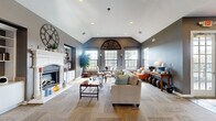Located in Maumelle, AR, this single-floor space offers a well-appointed layout combining a living room and kitchen into a cohesive, functional environment. The living room is the centerpiece of the space, defined by soaring vaulted ceilings with recessed lighting and neutral gray walls that create a sophisticated backdrop. A classic white fireplace with built-in shelving anchors one end of the room, while an array of large, grid-pane windows floods the interior with natural light and provides views of the surrounding outdoor greenery. A decorative arched wall ornament serves as a distinctive focal point above the window arrangement. The open layout comfortably accommodates multiple seating and dining areas, including a glass-top dining table and an inviting lounge arrangement. The kitchen features white cabinetry, a herringbone tile backsplash, stainless steel appliances, and slate tile flooring, offering a clean and practical workspace. A large window in the kitchen extends the sense of openness and connects the interior to outdoor views. Throughout the space, textured carpet and tile flooring complement the warm, welcoming ambiance of this thoughtfully designed environment.