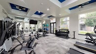 Located on Beechnut Street in Richmond, TX, this fitness center is a thoughtfully designed exercise facility that delivers a comprehensive workout environment. The single-floor space features a contemporary aesthetic defined by light gray carpeted flooring, crisp white walls, and a distinctive tray ceiling accented with bold navy blue panels and ceiling fans that enhance air circulation throughout.

Large windows line the walls on multiple sides, flooding the room with natural light and offering views of the surrounding outdoor amenities, including a pool area and landscaped grounds. Recessed lighting supplements the natural brightness, ensuring a well-lit atmosphere at any hour.

The exercise room is comprehensively outfitted with a full suite of fitness equipment. A row of treadmills and an elliptical trainer anchor the cardio zone, while a multi-station cable machine provides versatile strength-training options. A dumbbell rack with a wide range of free weights, stability balls, and a weight bench round out the strength and functional fitness offerings.

A standout design feature is the bold typographic mural spanning an entire wall, displaying motivational words such as "Inspire," "Stamina," "Energize," and "Dedication." Full-length mirrors on one wall amplify the sense of space and allow for form-checking during workouts. The overall ambiance is energetic, modern, and purpose-built for an effective fitness experience.