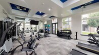 Located on Beechnut Street in Richmond, TX, this fitness center is a thoughtfully designed exercise facility that delivers a comprehensive workout environment. The single-floor space features a contemporary aesthetic defined by light gray carpeted flooring, crisp white walls, and a distinctive tray ceiling accented with bold navy blue panels and ceiling fans that enhance air circulation throughout.

Large windows line the walls on multiple sides, flooding the room with natural light and offering views of the surrounding outdoor amenities, including a pool area and landscaped grounds. Recessed lighting supplements the natural brightness, ensuring a well-lit atmosphere at any hour.

The exercise room is comprehensively outfitted with a full suite of fitness equipment. A row of treadmills and an elliptical trainer anchor the cardio zone, while a multi-station cable machine provides versatile strength-training options. A dumbbell rack with a wide range of free weights, stability balls, and a weight bench round out the strength and functional fitness offerings.

A standout design feature is the bold typographic mural spanning an entire wall, displaying motivational words such as "Inspire," "Stamina," "Energize," and "Dedication." Full-length mirrors on one wall amplify the sense of space and allow for form-checking during workouts. The overall ambiance is energetic, modern, and purpose-built for an effective fitness experience.