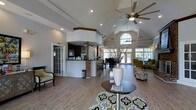 This single-story space is defined by its dramatic vaulted ceilings, which lend an airy, expansive feel throughout the open-concept interior. A large ceiling fan anchors the central living area, complementing the recessed lighting that illuminates the space evenly. The flooring throughout is a warm hardwood-style material that ties the areas together cohesively.

A curved bar-height counter with brick base detailing serves as a natural divider between the kitchen and main gathering area. The kitchen features dark cabinetry, under-cabinet track lighting, and bar stool seating, making it both functional and inviting. A glazed French door nearby adds architectural interest and provides access to an adjacent area.

The living area is comfortably furnished with patterned seating and a round accent table atop a bold geometric area rug. Decorative tall branches in a vase add a natural element to the décor. At the far end, a generous arched window flanked by additional windows allows abundant natural light to pour in, offering views of the surrounding greenery and enhancing the welcoming, open ambiance of the space.