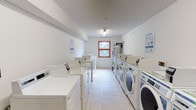 This shared laundry facility is a clean, efficiently organized space designed to serve multiple users with ease. The room is laid out in a linear corridor-style arrangement, with rows of top-loading washing machines lining one side and a combination of front-loading washers and dryers along the opposite wall. The machines are all modern, white-finished units equipped with clearly labeled controls, ensuring straightforward operation for users. Informational signage is posted on the walls, providing guidance on proper machine use and care instructions. The space is finished with neutral white walls and light-toned tile flooring, contributing to a bright and sanitary atmosphere. A single window with a warm wood-toned frame is positioned at the far end of the room, allowing natural light to filter in and providing a view of surrounding greenery. Overhead fluorescent lighting ensures the space remains well-lit throughout. A water heater is also visible at the rear of the room, supporting the facility's operational needs. The overall ambiance is practical and well-maintained, reflecting a facility that prioritizes cleanliness, functionality, and user convenience.