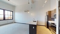 This single-floor unit showcases a contemporary loft-inspired design, blending industrial character with modern finishes throughout. The open-concept living and kitchen area is anchored by soaring ceilings that amplify the sense of space, complemented by polished concrete flooring and an exposed black structural column that nods to the building's industrial heritage. Overhead, a combination of pendant lighting and a track lighting system provides layered illumination across the living space. A wall-mounted mini-split unit ensures year-round climate control.

The kitchen is a standout feature, centered around a large island with a white quartz countertop and an undermount sink. Rich dark-toned cabinetry lines the walls, providing ample storage, while a full suite of stainless steel appliances — including a refrigerator, electric range, and built-in microwave — offers modern functionality.

The bedroom is a calm, carpeted retreat featuring whitewashed brick walls and large black-framed windows that frame open rooftop views and fill the room with natural light. A wall-mounted mini-split unit is also present here for independent climate control.

The bathroom is finished with warm bamboo-style hardwood flooring, a dark wood vanity with white countertop, a full-width mirror, and a separate toilet compartment, all framed by a sleek dark-framed door.
