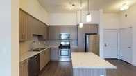 This single-floor space showcases a thoughtfully designed kitchen that serves as its centerpiece. The kitchen is characterized by soaring ceilings that amplify the sense of openness and volume. Rich, dark wood-grain cabinetry lines the walls, extending to near ceiling height to maximize storage capacity. A decorative geometric tile backsplash adds texture and visual interest between the upper and lower cabinetry. The countertops feature light-toned granite with subtle speckled detailing, offering both durability and elegance. A kitchen island with matching granite extends into the space, providing additional workspace and a natural gathering area. The full suite of stainless steel appliances includes a gas range, built-in microwave, refrigerator, and dishwasher, delivering a cohesive, professional-grade aesthetic. Overhead, a combination of pendant lighting and a ceiling-mounted track light system provides layered, adjustable illumination throughout the kitchen. The flooring consists of warm-toned hardwood planks that contrast effectively with the cabinetry. Overall, the space conveys a clean, contemporary ambiance with high-quality finishes throughout.