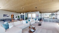 This sophisticated single-floor amenity space offers a generous, open-plan layout defined by a striking blend of modern industrial and contemporary design elements. Throughout the space, warm natural wood plank ceilings contrast beautifully with polished concrete-finish walls and large-format tile flooring, creating a refined yet welcoming atmosphere.

The main lounge area serves as the heart of the space, anchored by a dramatic concrete accent wall housing a mounted flat-screen television and a sleek linear fireplace. Generous sectional sofas in neutral tones are accented with teal throw pillows, complemented by solid wood coffee tables. Floor-to-ceiling glass windows and curtain walls wrap the perimeter, flooding the interior with abundant natural light and offering expansive outdoor views.

A dedicated billiards and lounge zone features rich leather club chairs, a pool table, and additional wall-mounted display screens, all framed by the same sweeping window walls. The full-service bar area is equipped with a dark granite-topped counter, stylish round stools, industrial pendant lighting, and a mirrored back bar with open shelving.

An entry foyer with elevator access, high-top bistro tables, and potted greenery provides a welcoming transition into the space. Every zone flows seamlessly into the next, reinforcing the open, connected, and upscale character of this exceptional amenity floor.