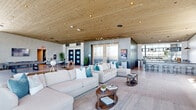 This sophisticated single-floor amenity space offers a generous, open-plan layout defined by a striking blend of modern industrial and contemporary design elements. Throughout the space, warm natural wood plank ceilings contrast beautifully with polished concrete-finish walls and large-format tile flooring, creating a refined yet welcoming atmosphere.

The main lounge area serves as the heart of the space, anchored by a dramatic concrete accent wall housing a mounted flat-screen television and a sleek linear fireplace. Generous sectional sofas in neutral tones are accented with teal throw pillows, complemented by solid wood coffee tables. Floor-to-ceiling glass windows and curtain walls wrap the perimeter, flooding the interior with abundant natural light and offering expansive outdoor views.

A dedicated billiards and lounge zone features rich leather club chairs, a pool table, and additional wall-mounted display screens, all framed by the same sweeping window walls. The full-service bar area is equipped with a dark granite-topped counter, stylish round stools, industrial pendant lighting, and a mirrored back bar with open shelving.

An entry foyer with elevator access, high-top bistro tables, and potted greenery provides a welcoming transition into the space. Every zone flows seamlessly into the next, reinforcing the open, connected, and upscale character of this exceptional amenity floor.
