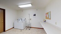 This single-floor shared laundry facility is designed with functionality and cleanliness in mind. The room features bright, even overhead fluorescent lighting that illuminates the entire space clearly, creating a practical and welcoming environment for daily use. The walls are finished in crisp white, contributing to the room's clean and organized appearance, while light-toned tile flooring adds durability and ease of maintenance. Two top-loading washing machines are positioned side by side along one wall, accompanied by a matching dryer, offering ample capacity for residents. A wire laundry cart on wheels provides convenient transport of laundry within the space. A plumbed utility hookup is available on the wall, indicating flexibility for additional appliances or equipment. A generous, continuous countertop runs along the adjacent wall, providing substantial surface area for folding and sorting laundry. A cork bulletin board is mounted on the wall, serving as a community communication hub for notices and announcements. The space is thoughtfully laid out to allow easy movement and access to all equipment, making it a highly practical shared amenity.