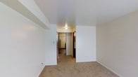 This single-floor space presents a straightforward and functional layout characterized by clean white walls and neutral-toned carpeting throughout. The main living area is open and unobstructed, offering a blank canvas that can accommodate a variety of furniture arrangements. A central hallway extends from the living space, connecting the various rooms within the unit and providing a clear sense of flow and accessibility.

The hallway is illuminated by a ceiling-mounted light fixture, ensuring adequate brightness through the passage. Dark-toned doors line the corridor, providing a subtle contrast against the white walls. At the end of the hallway, the kitchen is partially visible, featuring a white refrigerator and what appears to be additional cabinetry, suggesting a compact yet practical food preparation area.

The carpeting underfoot contributes a soft, warm quality to the interior, running consistently across the main living zone and hallway. White baseboards frame the perimeter of each room, adding a clean, finished appearance. The smooth white ceilings and minimal architectural detailing give the space a simple, utilitarian character, making it well-suited for practical, everyday living.
