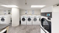 This shared laundry facility is organized across a single floor and divided into two functional areas designed to serve users efficiently and comfortably. The main laundry room is lined with rows of commercial-grade front-load washing machines and dryers positioned along the walls, providing ample capacity for high-volume use. A central folding table with a stone-finish countertop offers a convenient workspace for sorting and folding laundry. The room is finished with durable tile flooring in a grey stone aesthetic, white walls, and a drop ceiling with large flush-panel light fixtures that provide bright, even illumination throughout. A sliding glass door separates the laundry area from an adjacent lounge space, which features the same tile flooring and drop ceiling. The lounge is furnished with modern teal seating and accented by a decorative wall mural and a potted ornamental grass plant, creating a welcoming environment for waiting. A payment or card-reader station is visible near the entrance, indicating a card-operated system. Informational signage is posted throughout the laundry room for user guidance. The overall design is clean, functional, and well-maintained, making this facility a practical and user-friendly amenity.