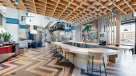 Located at 1010 E Orange St in Phoenix, Arizona, this thoughtfully designed amenity space delivers a bold and contemporary atmosphere that blends social, work, and lounge functions seamlessly. The space is anchored by a standout geometric coffered wood ceiling that stretches across a central zone, complemented by suspended blue pendant lights that add pops of color overhead. Soaring ceiling heights throughout create an open, airy feel, while large windows along the perimeter flood the interior with abundant natural light and offer views of the surrounding outdoor greenery. The flooring features an eye-catching chevron wood pattern that transitions into polished concrete, visually defining distinct zones within the open plan. A curved, white-topped bar with bar seating serves as a central social hub, accented by vibrant large-scale artwork on the adjacent wall. A comfortable lounge area is anchored by a circular patterned rug and rounded teal upholstered chairs arranged around a central coffee table, ideal for casual gatherings. Slatted wood partitions and built-in planters with lush greenery create soft separations between zones. A dedicated co-working area features long communal tables, bar-height seating, and colorful pendant lighting, while framed artwork and vertical slat screens contribute to the refined, design-forward aesthetic throughout.