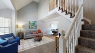 This two-story home opens into a spacious living area distinguished by dramatic vaulted ceilings that create an immediate sense of openness and volume. The living room features soft carpeting, neutral gray walls, and an open layout that flows seamlessly into the dining area, which is illuminated by a ceiling fan and receives natural light through sliding glass doors fitted with vertical blinds. A white-baluster staircase anchors the main level and leads to an upper floor, where an open overlook railing allows views down into the living space below.

The kitchen is outfitted with white cabinetry, granite-style countertops, a white subway tile backsplash, and black appliances including a range, over-the-range microwave, and refrigerator. Wood-style flooring grounds the kitchen space.

The bedrooms are carpeted and each feature ceiling fans, windows with blinds, and neutral walls. One bedroom includes a closet and ample room for furniture. A full bathroom serves the home with a granite-topped vanity, a tub-shower combination with white tile surround, and wood-style flooring, offering a clean, functional aesthetic throughout.