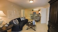 Located at 1201 Aragon Dr in Knightdale, NC, this single-floor residence offers a thoughtfully designed open-concept layout that seamlessly connects the living and kitchen areas. The living room is a welcoming, generously sized space anchored by plush carpeting and neutral wall tones that create a warm, inviting ambiance. A flush-mount ceiling fixture provides overhead lighting, complementing the soft, cozy atmosphere of the room. The space comfortably accommodates a full seating arrangement, making it ideal for both relaxing and entertaining. Adjacent to the living area, the kitchen features warm wood-toned cabinetry and a dark countertop breakfast bar with bar stool seating, offering a casual dining option and a natural transition point between the two spaces. The kitchen is well-equipped and benefits from the open sightlines to the living room, enhancing the social flow of the home. Tasteful framed wall art and decorative accents add a refined, classic touch throughout the space. Multiple doorways lead to additional rooms, suggesting a well-organized floor plan with comfortable private spaces. The overall design balances functionality and style, with consistent neutral tones and traditional detailing that give the home a timeless, polished character.