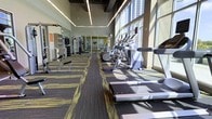 This fitness center is designed to offer a comprehensive workout experience within a thoughtfully arranged, single-level space. Floor-to-ceiling windows line one side of the room, flooding the interior with natural light and creating an open, energizing atmosphere. The generous ceiling height, accentuated by sleek linear pendant lighting and exposed structural beams, contributes to the spacious feel of the facility.

The cardio zone features a row of elliptical trainers and treadmills positioned along the windowed wall, allowing users to exercise with an abundance of natural light. On the opposite side, a dedicated strength training area is equipped with cable machines, free weights, and weight benches, complemented by large mirrors that visually expand the space and allow users to monitor their form. The flooring consists of patterned, multi-tonal carpet tiles in charcoal and yellow-green, adding a modern design element while providing cushioning underfoot.

The overall layout is organized and functional, with clear zones for both cardio and strength activities. The combination of modern finishes, ample lighting, and large windows creates a bright, motivating environment well-suited for a variety of fitness routines.