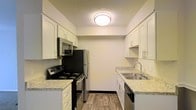 This single-floor space includes a well-appointed galley-style kitchen designed for efficient use of space. The kitchen features white cabinetry with sleek hardware lining both sides of the corridor-style layout, providing generous storage. Light granite-style countertops run along each wall, offering practical prep space. Stainless steel appliances include an electric range, over-the-range microwave, refrigerator, and dishwasher, lending a modern, cohesive look. A stainless steel sink with a contemporary faucet is positioned along one counter, complemented by a window above for added functionality. The wood-look plank flooring adds warmth and durability underfoot, while a flush-mount ceiling light provides even illumination throughout. The textured ceiling and neutral wall tones contribute to a clean, understated aesthetic. The galley layout maximizes efficiency, making excellent use of the available footprint. Overall, the kitchen presents a practical and updated environment well-suited for everyday cooking and meal preparation.
