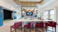 This single-floor community amenity space is thoughtfully designed with a modern coastal aesthetic, combining vibrant accent colors, natural wood tones, and crisp white finishes throughout. At the heart of the space is a large kitchen island topped with white quartz countertops, paired with rich burgundy upholstered bar seating. An eye-catching brass chandelier with globe pendants hangs above the island beneath a wood-paneled tray ceiling, adding warmth and sophistication. A mosaic tile feature wall serves as a striking backdrop behind the built-in sink and integrated appliances, with a mounted flat-screen TV completing the entertaining focal point. The adjacent lounge area is generously proportioned, featuring soaring ceilings with a coffered wood-inlay detail and linear pendant lighting. Deep burgundy and mauve sectional sofas and tufted blue accent chairs are arranged around a central coffee table, with multiple windows flooding the space with natural light and offering views of the surrounding landscape. A bold blue sliding barn door leads to a private golf simulator room, while glass double doors connect to a separate game room housing a full-size billiards table. Light wood-plank flooring flows seamlessly throughout, tying the distinct zones together into one cohesive, inviting communal retreat.