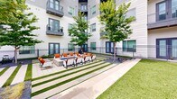 Located at 3700 Ross Ave in Dallas, TX, this contemporary residential community showcases a thoughtfully designed outdoor environment that emphasizes both aesthetics and functionality. The complex is defined by its modern multi-story architecture, featuring a combination of neutral stucco, gray paneling, and dark-framed windows, with private balconies visible throughout the upper floors.

At the heart of the community lies a generously sized resort-style swimming pool, surrounded by an expansive paver deck lined with rows of lounge chairs, offering ample space for relaxation. A shallow wading area provides additional versatility, while large floor-to-ceiling glass walls of an adjacent amenity building reflect the open sky above.

Beyond the pool, the outdoor amenity spaces continue with a landscaped courtyard featuring a central fire pit seating area appointed with orange cushioned chairs, framed by striped turf-and-paver pathways and mature shade trees. A dedicated outdoor kitchen and pergola structure provide an ideal setting for gatherings.

Thoughtfully maintained raised garden planters with wrought iron railings and accessible walkways connect the various zones throughout the community. A broad open lawn provides additional green space, while lush plantings of tropical palms, ornamental shrubs, and seasonal foliage contribute to the community's vibrant, well-maintained character.