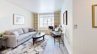 Located in the charming coastal city of Pacific Grove, California, this single-floor residence at 551 Gibson Ave offers a thoughtfully designed layout with a warm, neutral aesthetic throughout. The living area is a standout space, featuring built-in shelving units that flank a central window, flooding the room with natural light. Light hardwood-style flooring, a comfortable seating arrangement, and a built-in window seat with storage drawers create an inviting and functional atmosphere. The kitchen is efficiently laid out with light wood cabinetry, granite-style countertops, under-cabinet lighting, and white appliances, including a range and refrigerator, alongside a deep basin sink. The bedroom offers a quiet retreat with soft carpeting, a closet with built-in drawers, and a window dressed with layered curtains and a roller shade for adjustable light control. Two bathrooms serve the space, both featuring classic patterned tile flooring. The larger bathroom includes a wood-toned vanity with ample storage, a well-lit mirror, and accessibility features such as grab bars. The second bathroom is compact and similarly equipped with a shower-tub combination and safety grab bars. Overall, the space presents a clean, accessible, and comfortable living environment well-suited to Pacific Grove's relaxed coastal lifestyle.