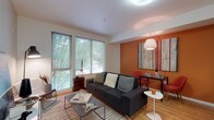 This well-designed single-floor residence combines modern aesthetics with warm, energetic color accents to create an inviting and functional living environment. The open-concept living and dining area serves as the heart of the space, anchored by large windows fitted with blinds that frame lush tree canopy views and flood the interior with natural light. Warm hardwood flooring runs throughout the main area, complementing a striking burnt-orange accent wall that adds depth and character. A contemporary pendant light fixture illuminates the dining nook, while the living area offers generous seating and media space. The kitchen is efficiently laid out with stainless steel appliances — including a refrigerator, electric range, and over-the-range microwave — paired with two-tone cabinetry and clean white countertops. The bedroom is a restful retreat featuring soft carpeting, dual bedside lighting, and a bold mustard-yellow accent wall that adds a lively touch to the otherwise neutral palette. The bathroom is cleanly finished with light flooring, a tub-shower combination, and warm wood-toned vanity cabinetry. Throughout the space, the cohesive use of color and contemporary furnishings creates a dynamic yet comfortable atmosphere.