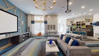 This stylish single-floor community lounge is designed with a contemporary aesthetic that balances comfort and functionality. The open-plan layout flows seamlessly from the main lounge area into a well-appointed kitchenette, creating a versatile space suited for both casual socializing and organized events.

The lounge area is anchored by a large sectional sofa in neutral grey, dressed with bold cobalt blue accent pillows and positioned atop a multi-toned striped area rug in grey, teal, and yellow. A geometric gold chandelier serves as a striking focal point overhead, complemented by additional globe pendant lighting throughout. A ceiling-mounted projector pairs with a large projection screen on a feature teal accent wall adorned with angular wood trim, adding a dynamic visual element to the space. Clerestory windows and full-height glazed entry doors allow generous natural light to fill the interior.

The kitchenette features dark cabinetry with granite countertops, a colorful mosaic tile backsplash, open display shelving, stainless steel appliances, and a spacious island with teal upholstered bar seating. A wall-mounted flat-screen television and a coffee station complete the amenity-rich kitchen zone. Wood-look flooring grounds the kitchenette, transitioning into the carpeted lounge area for a cohesive yet defined layout.