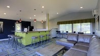 This single-floor space functions as a communal lounge and social hub, thoughtfully designed to encourage gathering and relaxation. The room is anchored by a bright lime-green bar counter fitted with a wooden countertop overhang and lined with sleek, adjustable bar stools—ideal for casual dining or conversation. Suspended above the counter, a pair of striking red pendant lights adds a bold pop of color and visual interest against the neutral gray ceiling and walls.

The kitchen or service area behind the counter features dark cabinetry and appliances, suggesting a functional preparation or refreshment zone. Recessed lighting throughout the ceiling provides consistent illumination across the space.

On the lounge side, a generously sized gray sectional sofa pairs with a rustic wooden coffee table, creating a comfortable seating arrangement suited for unwinding. A patterned area rug in muted blues and greens ties the two zones together, adding texture underfoot.

Large windows along one wall, dressed with olive-toned Roman shades and vertical blinds, allow natural light to filter in while offering a view of greenery outside. Additional high-top seating near the windows provides a secondary gathering spot, enhancing the open, versatile feel of the room.