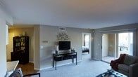 This single-floor residence features a thoughtfully laid-out living area finished with plush wall-to-wall carpeting and neutral gray-toned walls that create a calm, understated atmosphere throughout. The main living space is open and functional, centered around a sleek black media console topped with a flat-screen television and flanked by decorative candle holders, with a branch-motif metal wall accent adding an artistic touch above. Overhead, smooth white ceilings with a centrally located HVAC vent contribute to the clean, modern aesthetic. Natural light filters in through a glass-paneled exterior door situated along the far wall, brightening the space and providing direct outdoor access. A doorway visible from the living area leads directly into a bedroom, which features its own window allowing additional natural light and is furnished with a low-profile bed. The transition between rooms is seamless, with consistent neutral tones and white trim unifying the interior. Partial hardwood or hard-surface flooring is visible near the entryway, contrasting with the carpeted living zone. The overall ambiance is clean, neutral, and move-in ready.