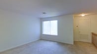 This single-story space presents a clean and understated interior highlighted by a well-proportioned room with soft, neutral-toned carpet underfoot. The walls are painted in a crisp white finish, contributing to an open and airy atmosphere throughout. A large window fitted with vertical blinds serves as the room's focal point, allowing generous amounts of natural light to fill the space and creating a bright, welcoming environment.

The textured ceiling adds a subtle architectural detail, while a centrally located HVAC vent ensures consistent climate control. Baseboards line the perimeter of the room, providing a polished transition between the walls and carpeted floor. Electrical outlets are thoughtfully positioned along the walls, offering practical functionality.

A doorway visible along one wall suggests additional rooms or spaces beyond, indicating a functional and connected layout. The overall ambiance of the space is calm and neutral, offering a versatile canvas well-suited to a variety of furnishing styles and uses. The simplicity of the design allows the natural light and clean lines to remain the defining characteristics of the interior.
