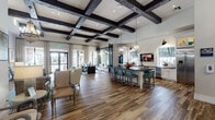 This thoughtfully designed single-floor community space blends rustic warmth with contemporary sophistication. The welcoming entrance foyer sets the tone immediately, featuring soaring ceilings, rich wood-look flooring, and a trio of striking geometric chandelier fixtures that cast a warm, inviting glow throughout.

The expansive main gathering area is defined by dramatic coffered ceilings with bold dark wood beams, recessed lighting, and generous natural light streaming through floor-to-ceiling windows dressed with plaid drapery. A teal-tiled fireplace anchors a comfortable lounge seating arrangement, creating an intimate focal point within the open layout. Nearby, a fully equipped kitchen area showcases white shaker cabinetry with dark hardware, dark countertops, stainless steel appliances including a full-size refrigerator and dishwasher, and a large central island with teal upholstered bar seating — ideal for gatherings and casual dining.

Exposed brick accents and dark structural columns add character and depth, while pendant globe lighting enhances the stylish, open-concept flow between the lounge and kitchen zones.

A dedicated screening room offers a private entertainment experience, outfitted with rows of plush leather reclining chairs with built-in cup holders, a ceiling-mounted projector, and floor-to-ceiling textured wallcovering that contributes to excellent acoustics and a true cinematic ambiance.