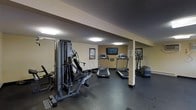 This single-level fitness room provides a functional workout environment designed to meet essential exercise needs. The space features dark rubber flooring, which offers durability and shock absorption suited for gym use. A multi-station cable and weight machine anchors one side of the room, accompanied by a large wall mirror that enhances the sense of space and allows users to monitor their form. Two treadmills are positioned centrally, facing a wall-mounted flat-screen television, making cardio sessions more engaging. An elliptical trainer is also available on the opposite side, rounding out the cardio equipment selection. Recessed fluorescent ceiling lighting provides consistent illumination throughout the room. The walls are finished in neutral tones, contributing to a clean and practical atmosphere. A structural support column is integrated into the layout, and informational fitness charts are posted on the walls for user reference. Overall, the fitness room is a straightforward, well-equipped space that prioritizes functionality and convenience for everyday workouts.
