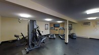 This single-level fitness room provides a functional workout environment designed to meet essential exercise needs. The space features dark rubber flooring, which offers durability and shock absorption suited for gym use. A multi-station cable and weight machine anchors one side of the room, accompanied by a large wall mirror that enhances the sense of space and allows users to monitor their form. Two treadmills are positioned centrally, facing a wall-mounted flat-screen television, making cardio sessions more engaging. An elliptical trainer is also available on the opposite side, rounding out the cardio equipment selection. Recessed fluorescent ceiling lighting provides consistent illumination throughout the room. The walls are finished in neutral tones, contributing to a clean and practical atmosphere. A structural support column is integrated into the layout, and informational fitness charts are posted on the walls for user reference. Overall, the fitness room is a straightforward, well-equipped space that prioritizes functionality and convenience for everyday workouts.