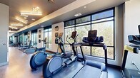 This contemporary fitness center offers a thoughtfully designed, single-level layout that accommodates a comprehensive range of workout activities. The space is anchored by a row of large, floor-to-ceiling windows along one wall, flooding the interior with natural light and providing serene, tree-lined views that create an inviting atmosphere for exercise.

The cardio zone features multiple treadmills and elliptical machines positioned directly in front of the expansive windows, allowing users to enjoy the outdoor scenery during their workouts. Adjacent to the cardio area, a cable machine and dumbbell rack provide a dedicated strength training section, complemented by a wall-mounted rack of kettlebells suited for functional fitness routines.

A full-length mirrored wall defines a flexible open floor area ideal for stretching, yoga, or mat-based exercises. Stability balls and additional accessories are visible throughout, adding to the versatility of the space. A small amenity station with organized storage is centrally located for convenience.

The design aesthetic is modern and clean, with warm wood-toned flooring, a two-tone color scheme of white and dark gray walls, and recessed lighting that ensures the space is well-lit throughout. The overall ambiance is professional, bright, and energizing — well-suited for a comprehensive fitness experience.