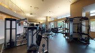 This expansive fitness center is thoughtfully designed across several distinct training zones, offering a comprehensive workout experience for users of all fitness levels. The main strength and cardio area features generous ceiling heights, with an open mezzanine-level detail adorned with warm golden vertical accents and sheer panels that lend an upscale, modern ambiance. Large windows along the perimeter flood the space with natural light and provide views of surrounding greenery, creating an inviting and energizing atmosphere. This zone is outfitted with a full lineup of Life Fitness machines, including cable systems, chest press, seated row, and multi-use strength stations, complemented by a rack of dumbbells and a dedicated flat bench. Cardio equipment includes ellipticals, recumbent bikes, and treadmills, several with integrated screens. A separate functional training studio features rich wood-plank flooring, a striking slatted wood ceiling with skylights, and a wall-mounted flat-screen TV. This room is equipped with spin bikes, stability balls, battle ropes, a rowing machine, kettlebells, and a barbell — ideal for group classes or high-intensity training. Throughout the facility, rubber flooring, mirrored walls, and accent lighting contribute to a polished, professional-grade environment designed to support a wide range of training goals.