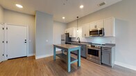 Located at 750 E Broad St in Columbus, Ohio, this single-floor unit offers a clean, modern aesthetic with a thoughtfully designed open layout. The kitchen serves as a central highlight, featuring white upper cabinetry, gray lower cabinets, a subway tile backsplash, dark countertops, and a full suite of stainless steel appliances including a refrigerator, range, microwave, and dishwasher. Pendant lighting and recessed fixtures illuminate the space, while a freestanding island provides additional prep and dining space. Durable wood-look flooring runs throughout the main living and kitchen areas, contributing to a cohesive, contemporary feel. The open-concept design flows seamlessly from the kitchen into the living area, which benefits from natural light streaming through vertical blind-covered sliding glass doors. The bedroom is finished with soft carpeting and features a large window with exterior views, offering a bright and comfortable retreat. The bathroom is accented with a bold teal feature wall, light wood-look tile flooring, and black hardware details, adding a distinctive touch. Built-in shelving provides convenient storage. The unit's neutral gray color palette, clean white trim, and modern fixtures create a polished and inviting atmosphere throughout.